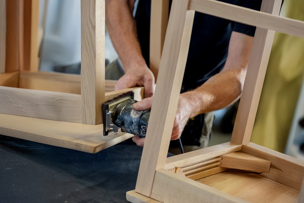 A craftsman sands the wooden frame of a chair with an orbital sander during the production process The image shows woodworking and artisanal furniture - Wurth Louis and Company A craftsman sands the wooden frame of a chair with an orbital sander during the production process The image shows woodworking and artisanal furniture